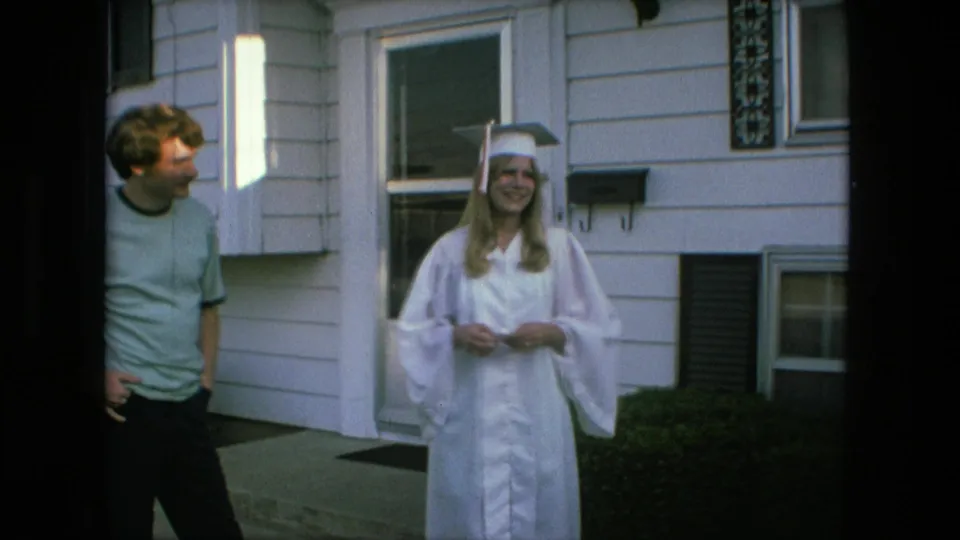 In Fort Wayne, Indiana in 1976, a young woman in a white graduation cap and gown smiles outside a suburban house while a young man in a pale T-shirt stands nearby.