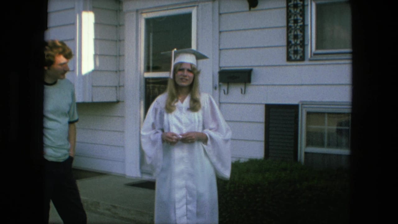 Graduate in white cap and gown faces the camera outside a suburban house.