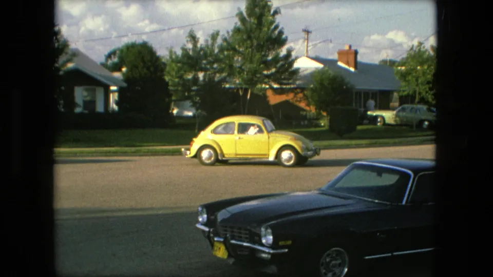 A Fort Wayne residential street in 1976 shows a yellow Volkswagen Beetle passing or parked near the curb with a dark coupe in the foreground and single-story houses behind.