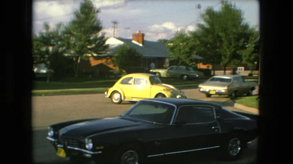 The 1976 Fort Wayne street view continues with a dark coupe in the foreground, a yellow Volkswagen Beetle across the road, and houses and trees in the background.