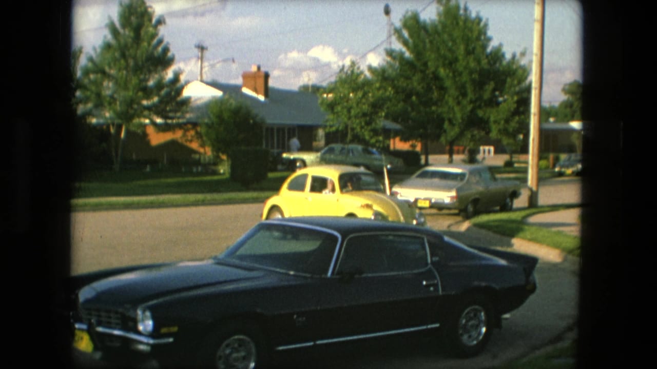 A yellow Volkswagen Beetle passes near a dark Chevrolet Camaro on a Fort Wayne suburban street in 1976.