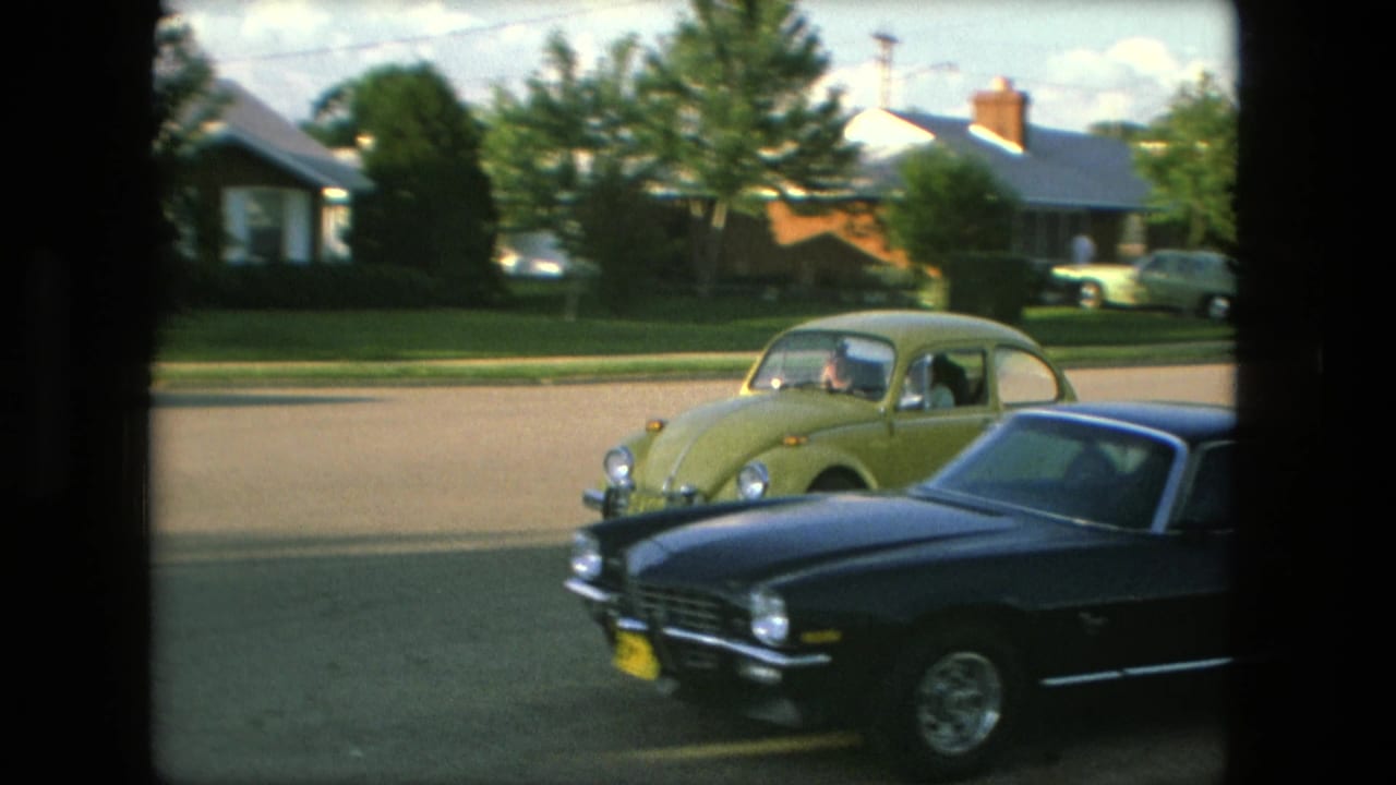 The yellow Beetle moves beside a dark Chevrolet Camaro with Fort Wayne houses and lawns in the background.