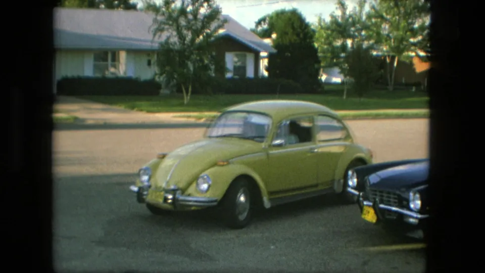 A yellow Volkswagen Beetle rolls past the front of a dark Chevrolet Camaro on a 1976 Fort Wayne residential street.