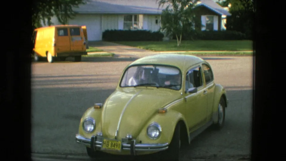 The yellow Beetle fills the foreground while an orange van and residential homes sit across the Fort Wayne street.