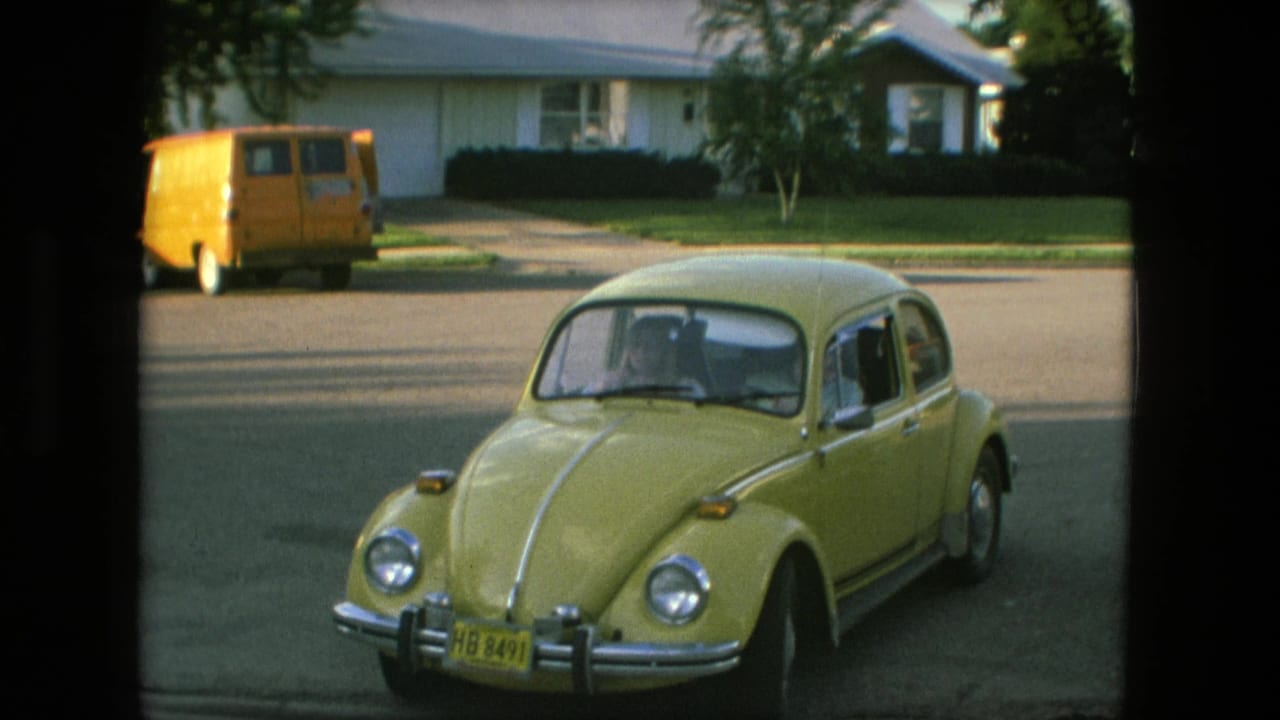 Yellow Volkswagen Beetle in a Fort Wayne suburban residential scene, with lawns, houses, and a parked yellow van visible beyond it.