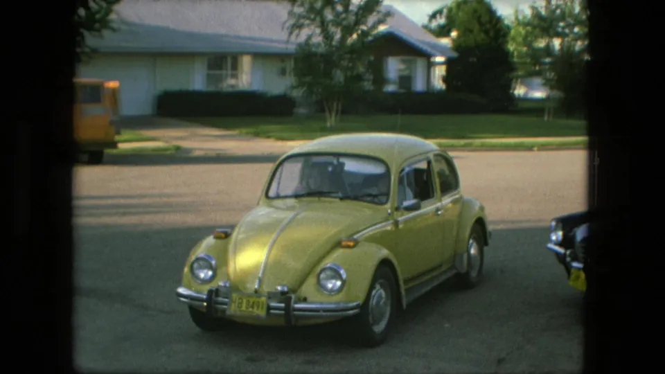Yellow Volkswagen Beetle pulls toward the street in a 1976 Fort Wayne neighborhood, with a parked yellow van and ranch-style homes behind it.