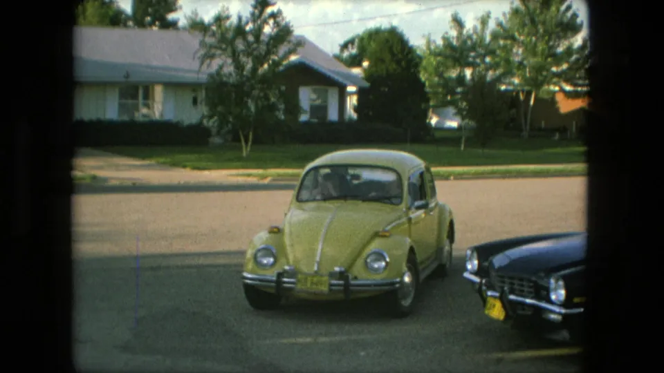 The yellow Beetle turns onto the street in Fort Wayne, with a dark car at the right edge and suburban houses across the road.