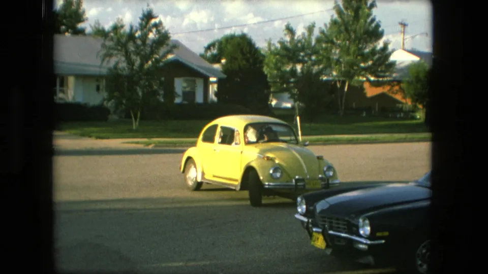 Yellow Volkswagen Beetle drives along a Fort Wayne residential street, with a dark parked car foregrounded and houses and lawns behind it.