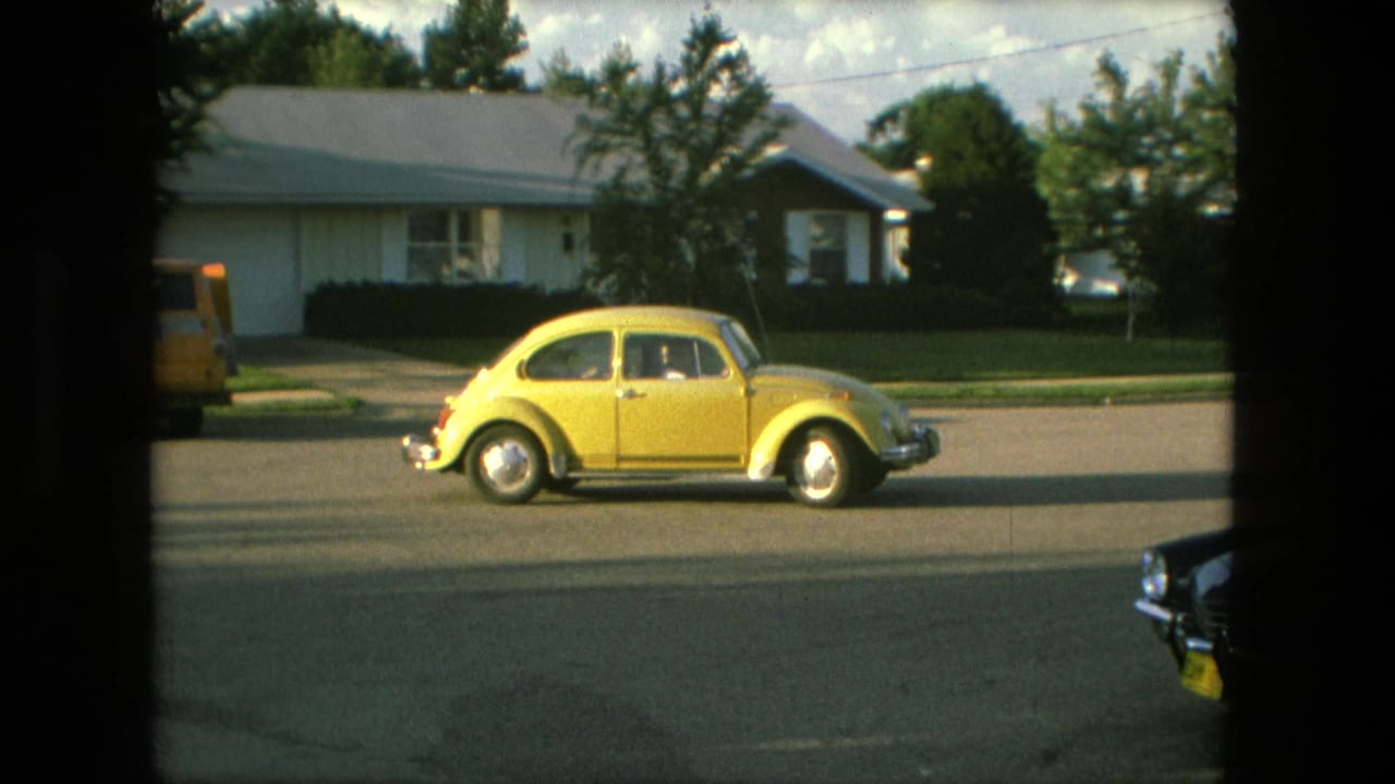 Yellow Volkswagen Beetle passing a house, with a dark car partly visible at right.