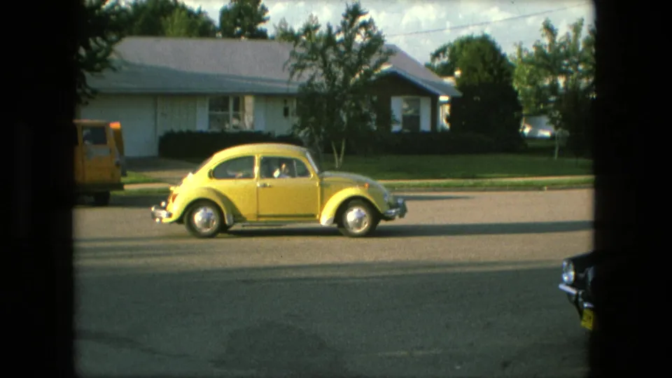 Yellow Volkswagen Beetle seen broadside on a Fort Wayne residential street, with houses and trees in the background.