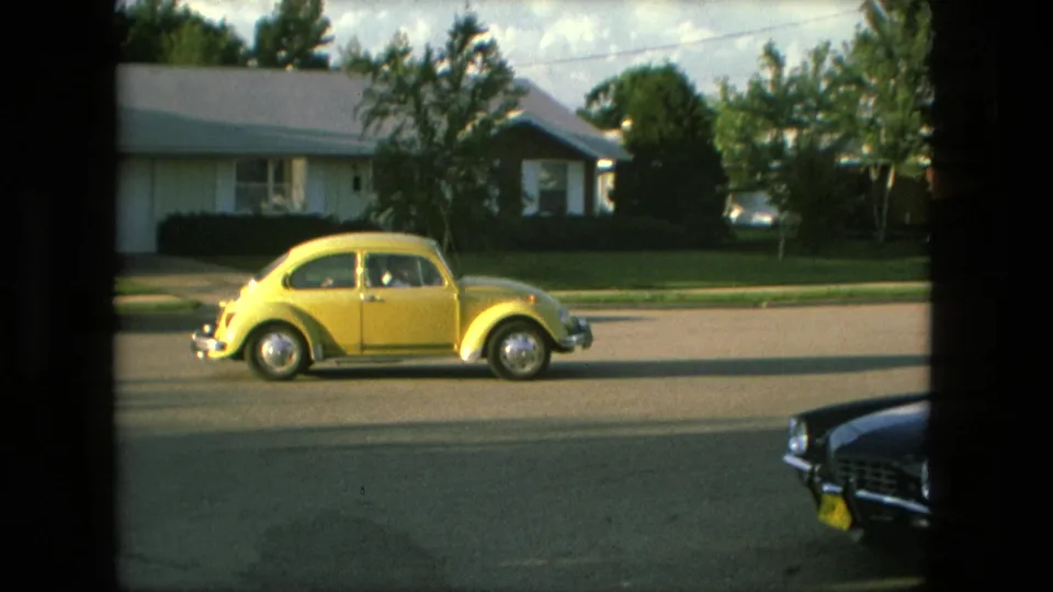 Yellow Volkswagen Beetle passes through a Fort Wayne neighborhood, partly framed by a dark foreground car.