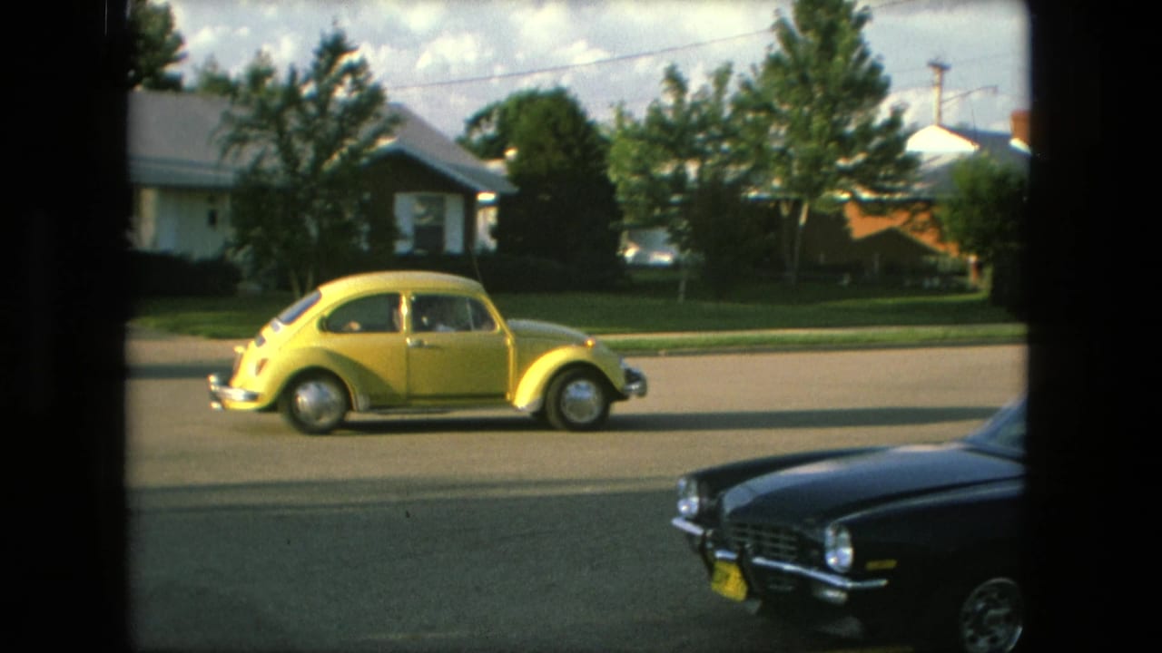 A yellow Volkswagen Beetle drives along a residential street in Fort Wayne, Indiana, in 1976, seen through a window frame with a dark coupe parked or passing in the foreground.