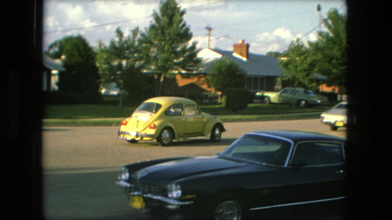 The yellow Volkswagen Beetle continues along the Fort Wayne residential street, with a dark coupe across the lower foreground and houses, lawns, and trees beyond.