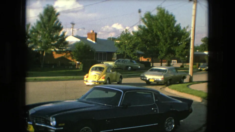 The Beetle approaches or passes a residential intersection in Fort Wayne, with a silver car, parked vehicles, lawns, utility pole, and a dark coupe visible through the window framing.