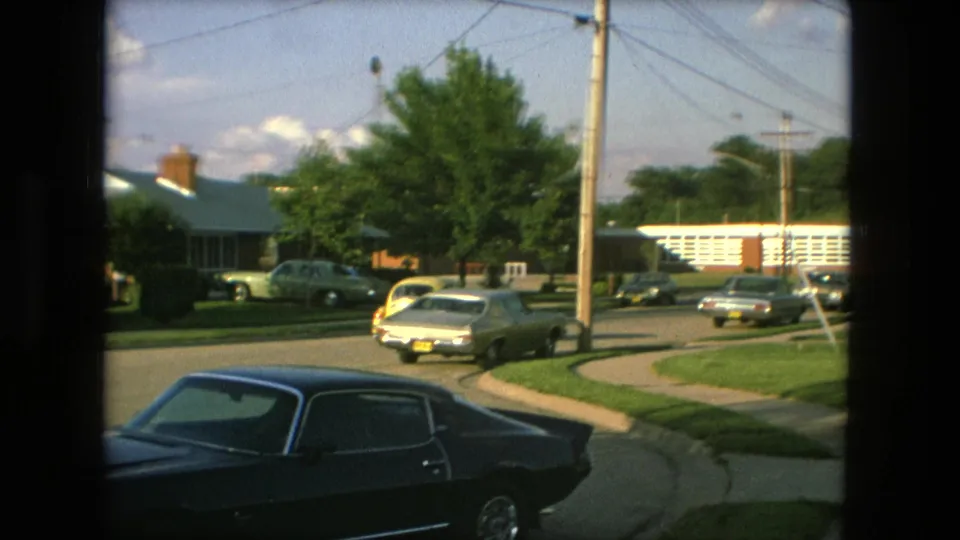 At the end of the clip, the Fort Wayne residential intersection shows the dark foreground coupe, parked cars, utility poles, sidewalks, lawns, and mid-century homes as the Beetle has mostly left the frame.
