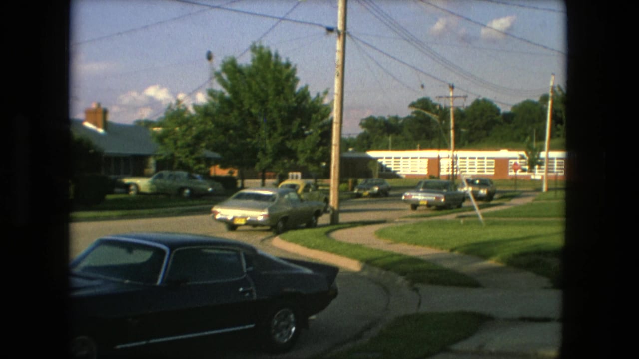 1976 Fort Wayne home-movie frame showing a suburban street with classic cars, sidewalks, utility poles, green lawns, and a low brick building in the background.