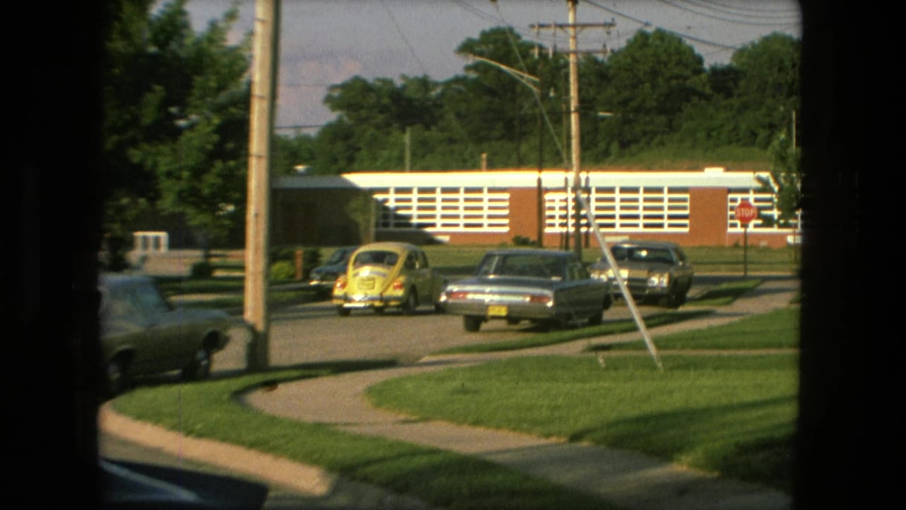 Yellow Beetle and sedans on a quiet street beside lawns and a brick building.