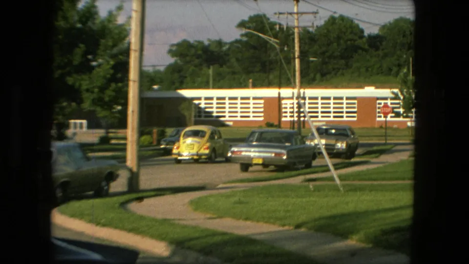 1976 Fort Wayne frame with a yellow Beetle, parked sedans, stop sign, utility poles, sidewalks, and a brick school-like building across the street.