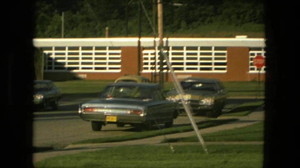 1976 Fort Wayne home-movie frame focused on classic sedans, a stop sign, utility pole, green lawn, and the white-windowed brick building in the background.