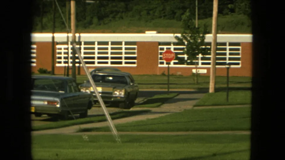 1976 Fort Wayne frame showing parked classic cars, a stop sign, utility poles, sidewalks, trimmed lawns, and a long brick building with white window panels.