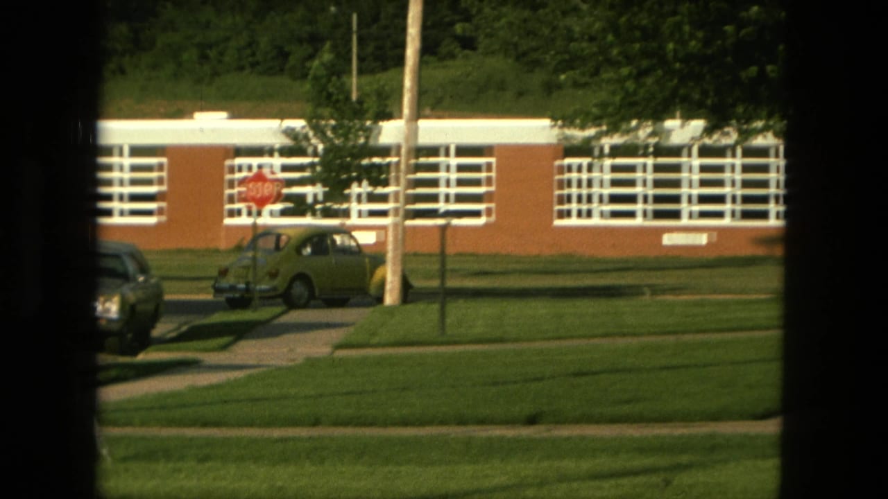 1976 Fort Wayne street view with a yellow Volkswagen Beetle, stop sign, lawn, utility pole, and brick school building in the background.