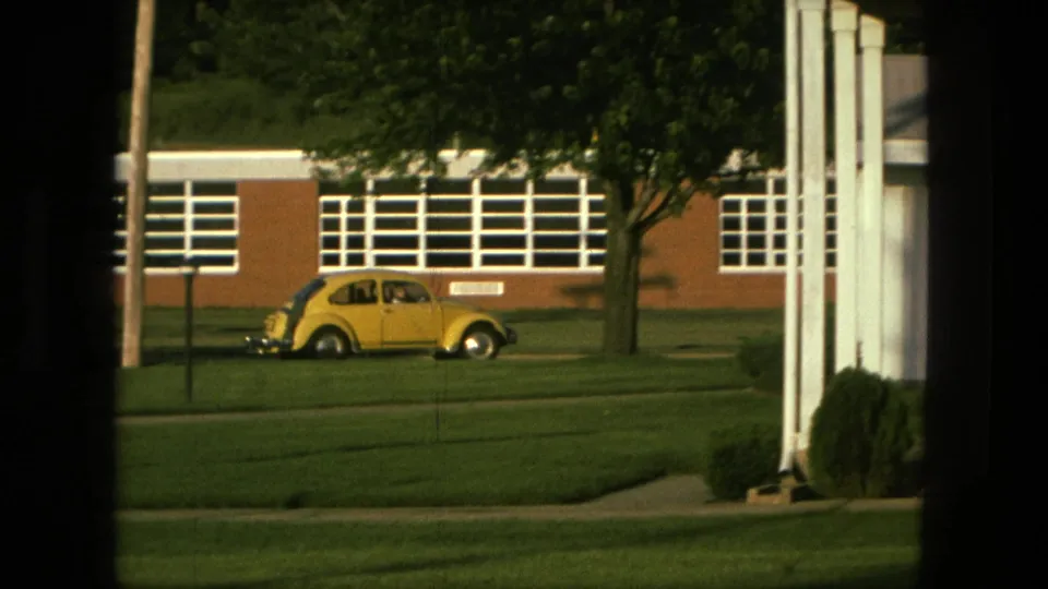 Home-movie frame showing a yellow Volkswagen Beetle along a green lawn, with a brick school building, white window bands, and a tree behind it.