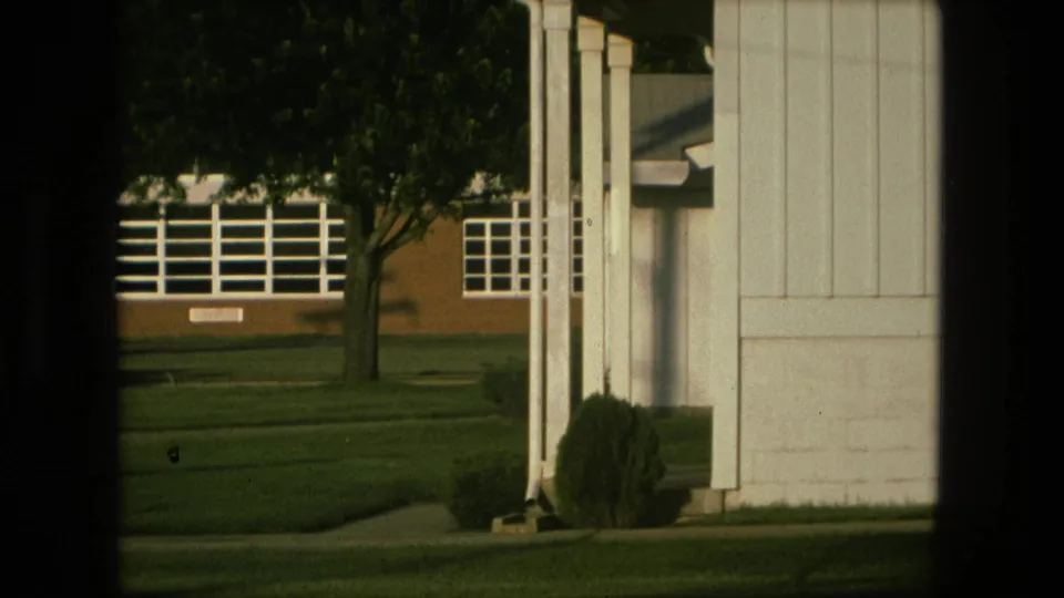 Fort Wayne home-movie frame with white house siding and shrubs in the foreground, facing a lawn, tree, and brick school across the street.