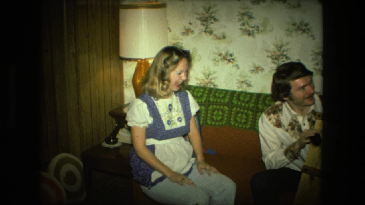 Woman in a blue and white dress sits laughing on a couch beside a man in a patterned shirt.