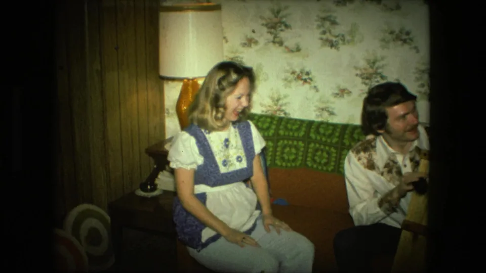 In a 1976 Fort Wayne home interior, a laughing woman in a blue and white dress sits on a couch while a man looks toward her.