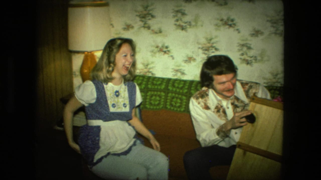 Laughing woman sits on a couch while a man examines a small wooden item nearby.