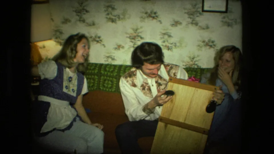 Three adults in a 1976 Fort Wayne living room share a light moment around a small wooden object near the couch.