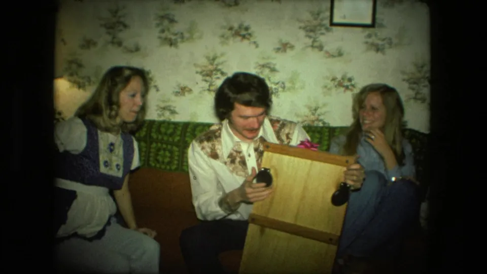 In the Fort Wayne home-movie frame, a man holds a small wooden wheeled object as two women watch from the couch.