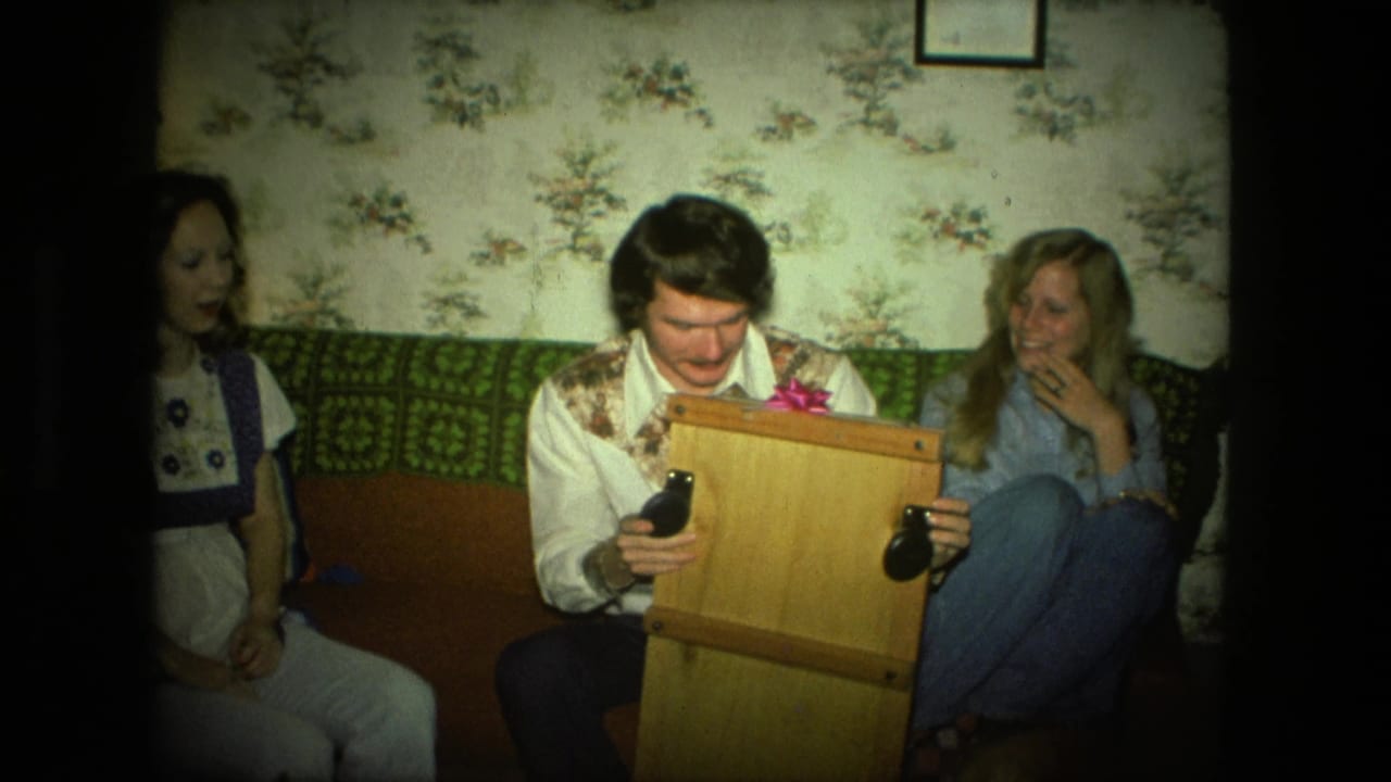 Man sits on a couch holding a wooden box while two women watch in a floral-wallpapered room.