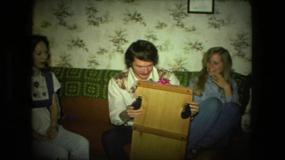 In a 1976 Fort Wayne home interior, a seated man holds a wooden gift box on a couch as two women look on.