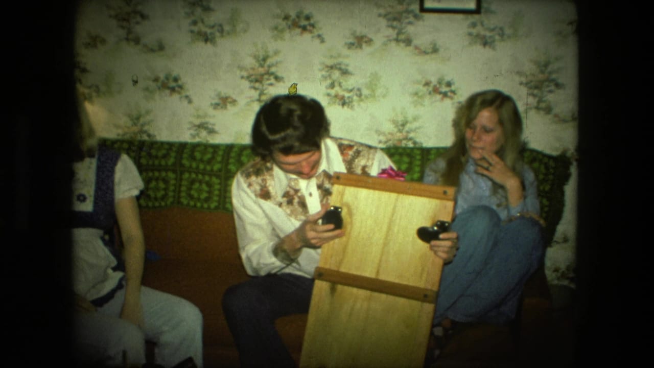 The man studies a small black handheld object beside the wooden gift box as a woman watches from the couch in the Fort Wayne home.