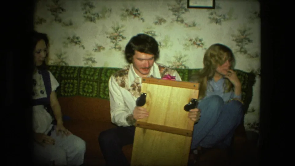A seated man in a patterned shirt holds the wooden present upright while two women sit nearby in a 1970s Fort Wayne living room.