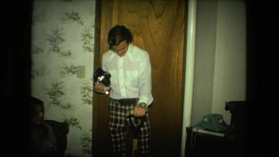 The man stands by a wooden door in the private Fort Wayne interior, holding a dark handheld object and strap while looking down.