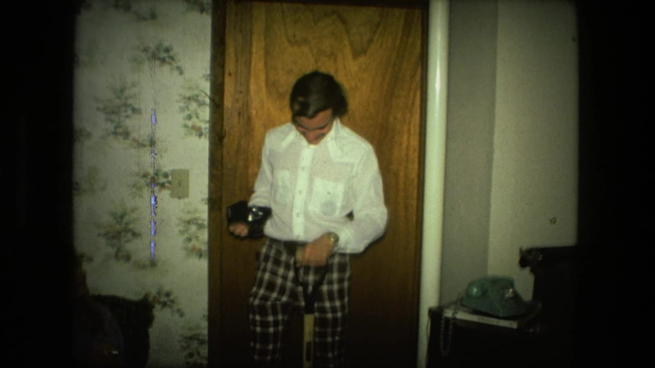 Man in a white shirt and plaid pants looks down while holding a camera near a wood door.