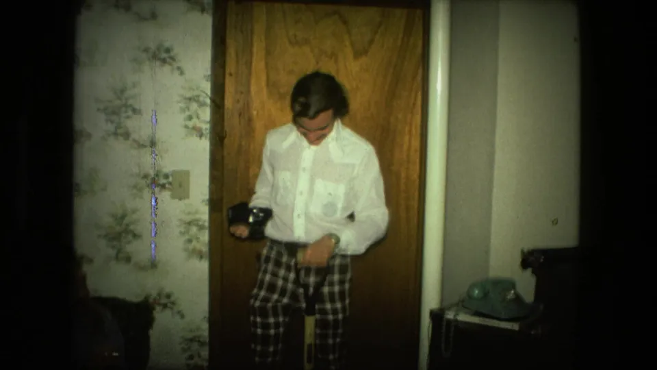 In a 1976 Fort Wayne home interior, a man in plaid pants looks down at a small camera beside floral wallpaper and a wood door.