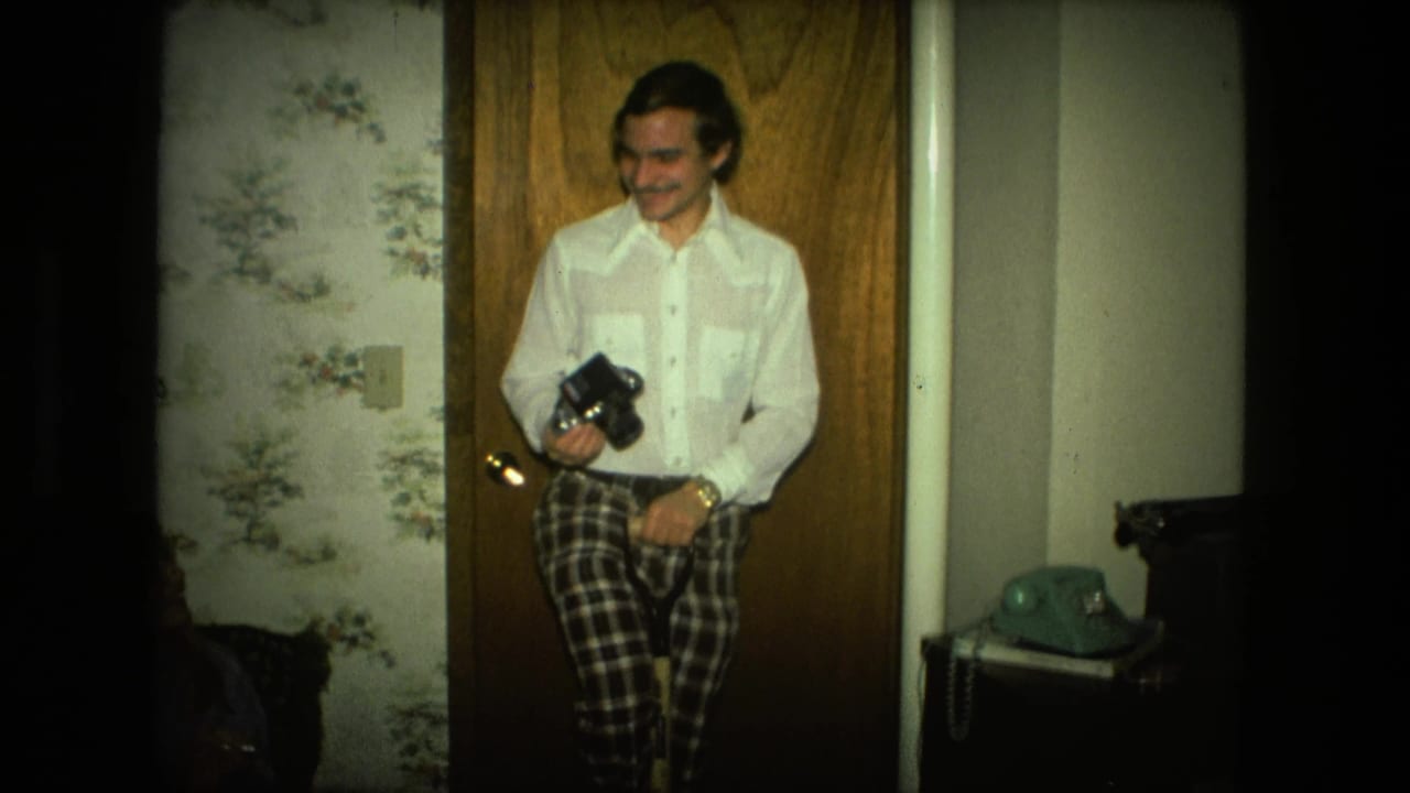 A smiling man stands in a 1976 Fort Wayne interior holding a camera, with floral wallpaper and a green rotary phone nearby.