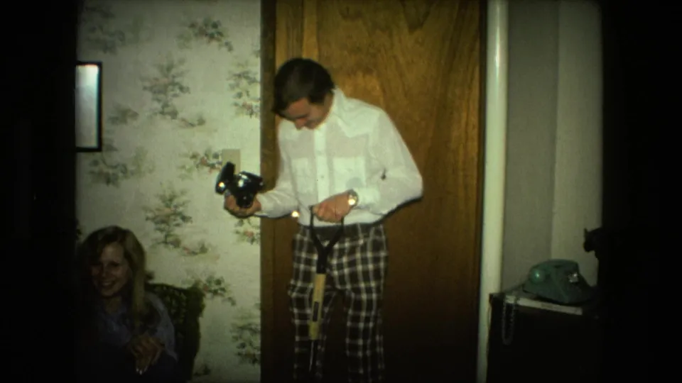 In the Fort Wayne home interior, the man adjusts or holds a shovel handle and camera while a seated woman smiles nearby.