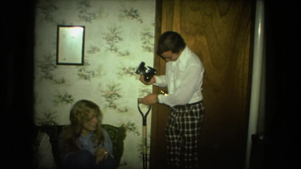 The man stands by the door with a camera and shovel handle while a smiling woman sits in the 1976 Fort Wayne home interior.