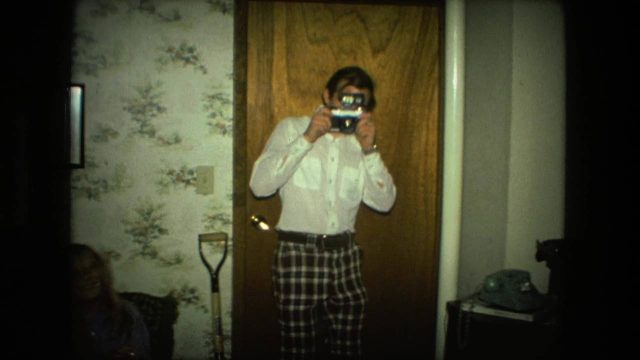 A man in a white shirt and plaid pants pretends to photograph someone in a 1976 Fort Wayne living room with a rotary phone nearby.