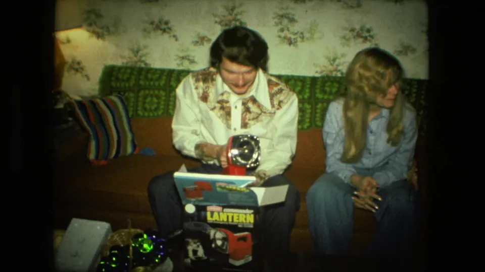 A seated man opens a lantern box beside a woman on a couch in a Fort Wayne home interior, with 1970s wallpaper and cushions visible.