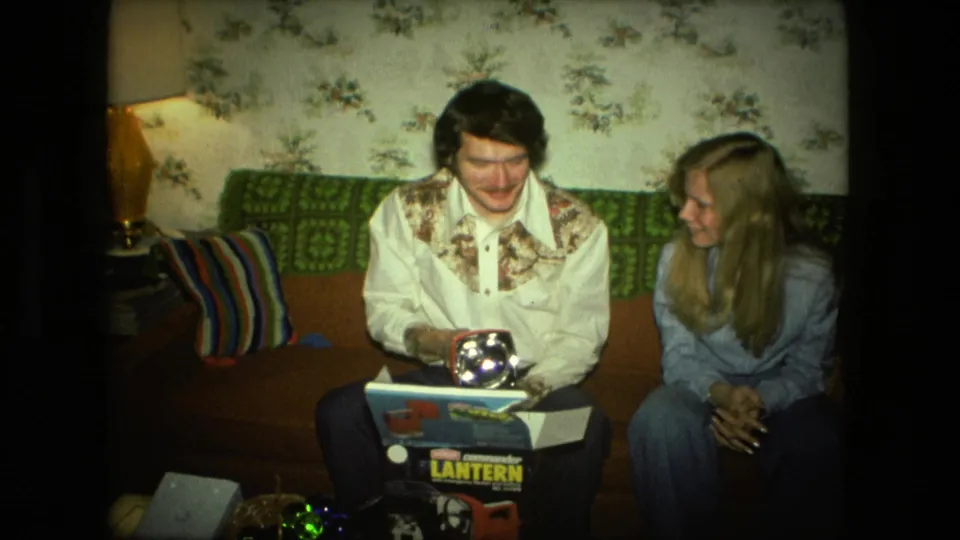 In the Fort Wayne living room, a man holds a shiny lantern above its box as a woman sits beside him on the couch.