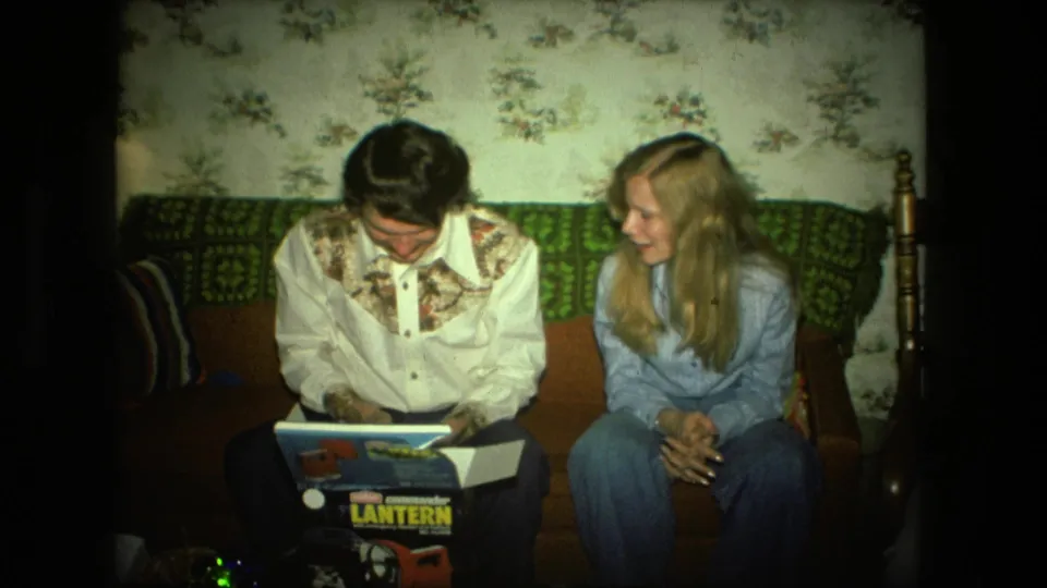 A 1976 Fort Wayne living room scene shows a young man inspecting a Coleman lantern box while a blonde woman leans in with interest.