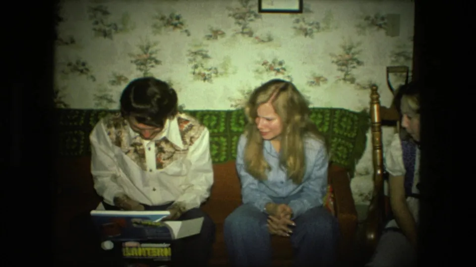 In the Fort Wayne home interior, the lantern box sits on the young man's lap while a blonde woman watches and a second woman is partly visible.