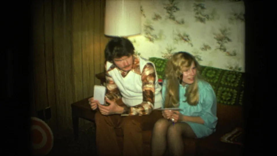 1976 Fort Wayne home-movie frame with a man and woman seated on a patterned couch in a Christmas living-room setting.