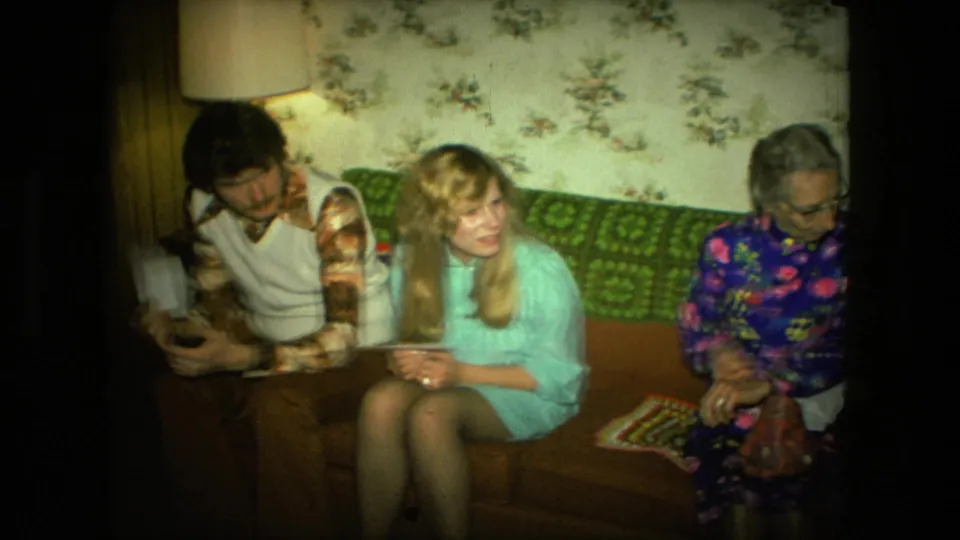 1976 Fort Wayne frame showing three adults on a couch in a private living room with floral wallpaper, wood paneling, and holiday gathering context.