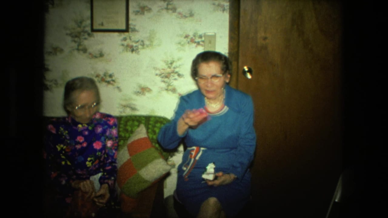 Two elderly women sit on a couch in a floral-wallpapered living room while one holds small craft pieces.